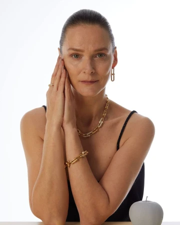 Close-up portrait of a mature woman wearing gold link jewelry and a black top against a white seamless background.