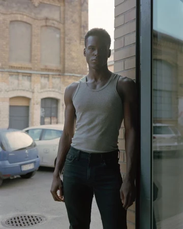 Young adult male with dark skin poses outdoors in a grey tank top and black jeans next to a brick wall and reflecting glass window.