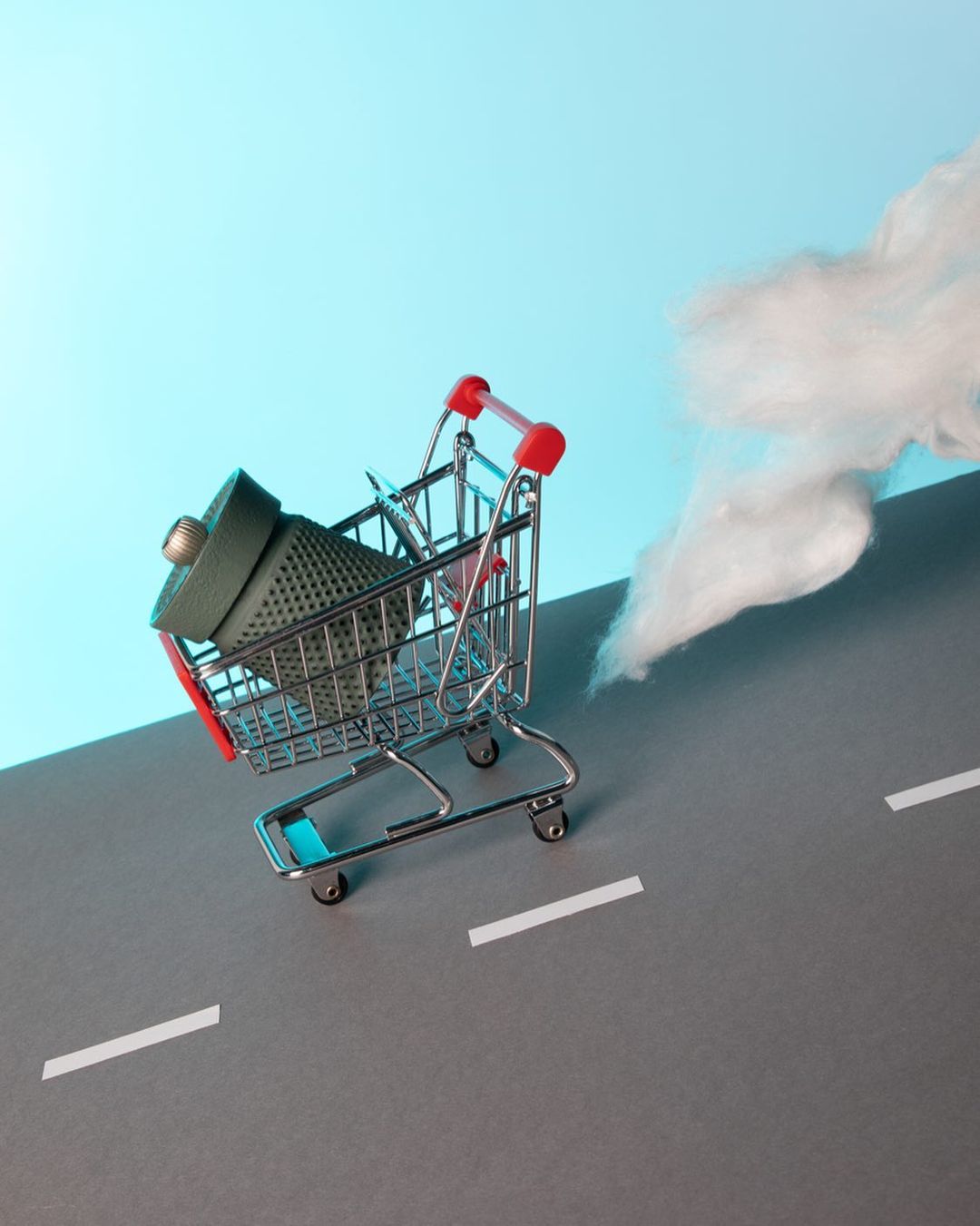 A miniature shopping cart containing a green pin cushion, with a white cloud-like plume behind it on a blue and gray background.