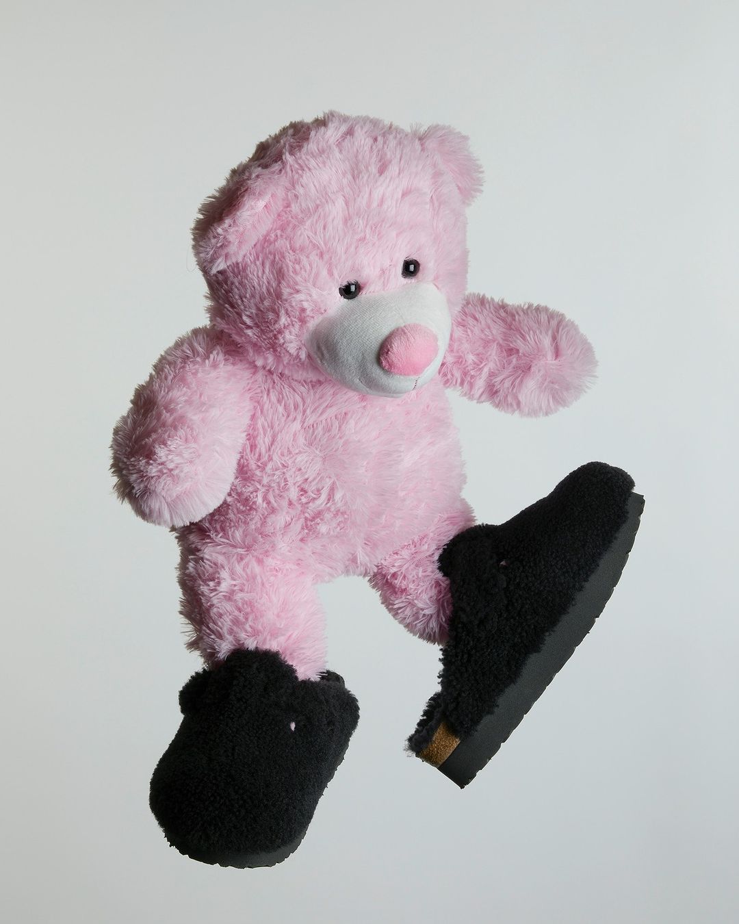 Pink teddy bear wearing black furry slippers and floating against a pale gray background in a studio product shot.