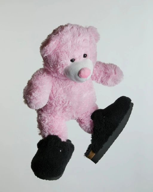 Pink teddy bear wearing black furry slippers and floating against a pale gray background in a studio product shot.