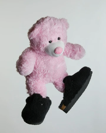 Pink teddy bear wearing black furry slippers and floating against a pale gray background in a studio product shot.