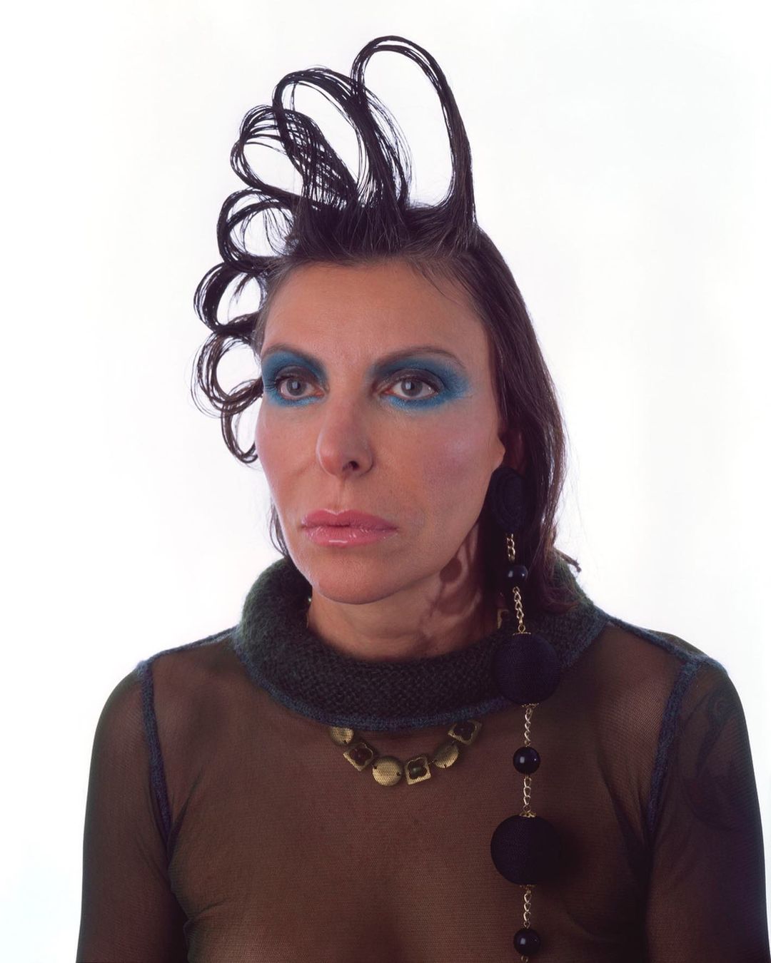 Portrait of an adult female with distinctive hair and makeup, adorned with statement jewelry, set against a bright white backdrop.