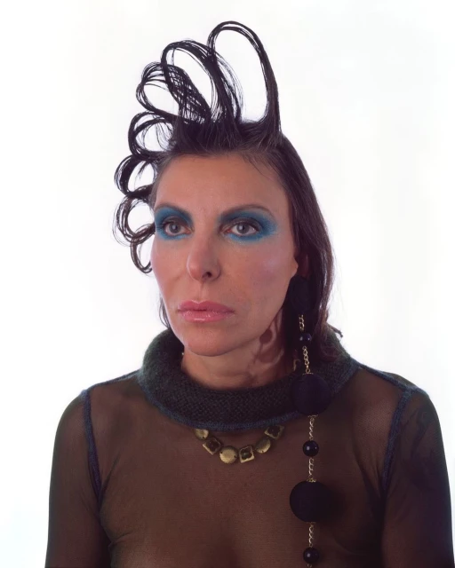 Portrait of an adult female with distinctive hair and makeup, adorned with statement jewelry, set against a bright white backdrop.