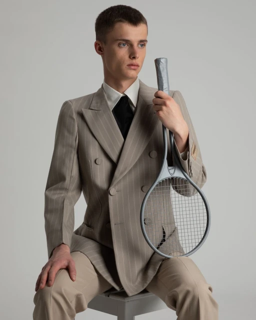 Young man in a pinstripe suit sits holding a vintage tennis racket in a studio, looking off to the side, with a neutral backdrop.