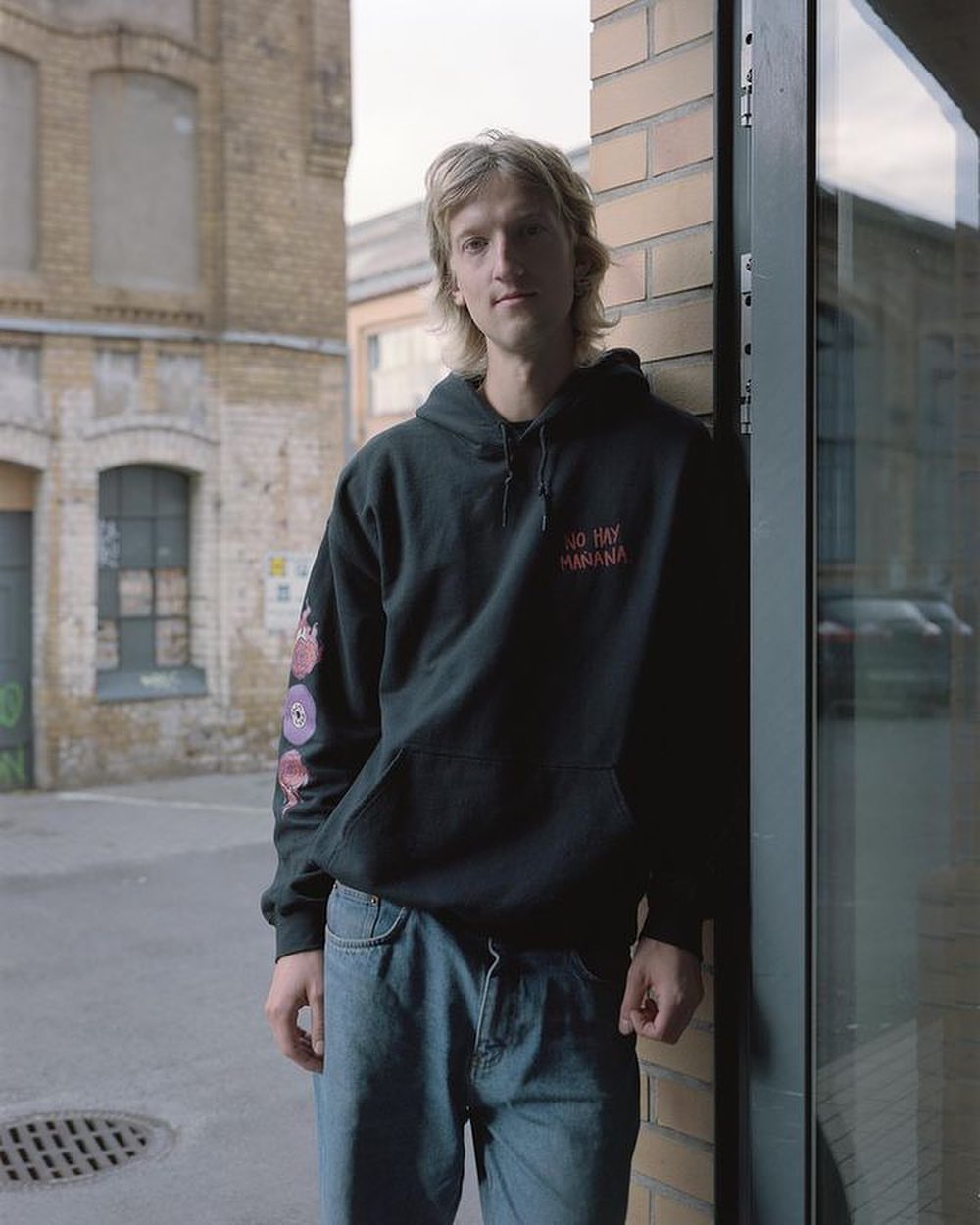 Adult male in black hoodie stands against a brick wall outdoors, showcasing a casual and urban style against an industrial backdrop.