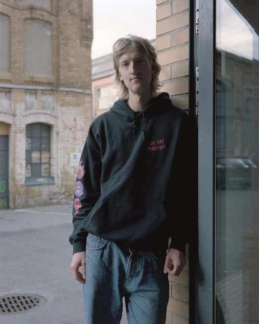 Adult male in black hoodie stands against a brick wall outdoors, showcasing a casual and urban style against an industrial backdrop.
