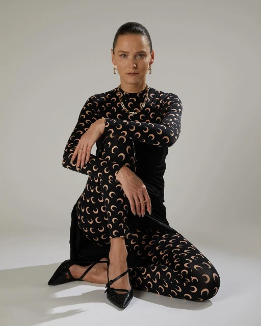 Portrait of a woman in a printed black dress posing in a photo studio. She is wearing gold jewelry and has her hair pulled back.