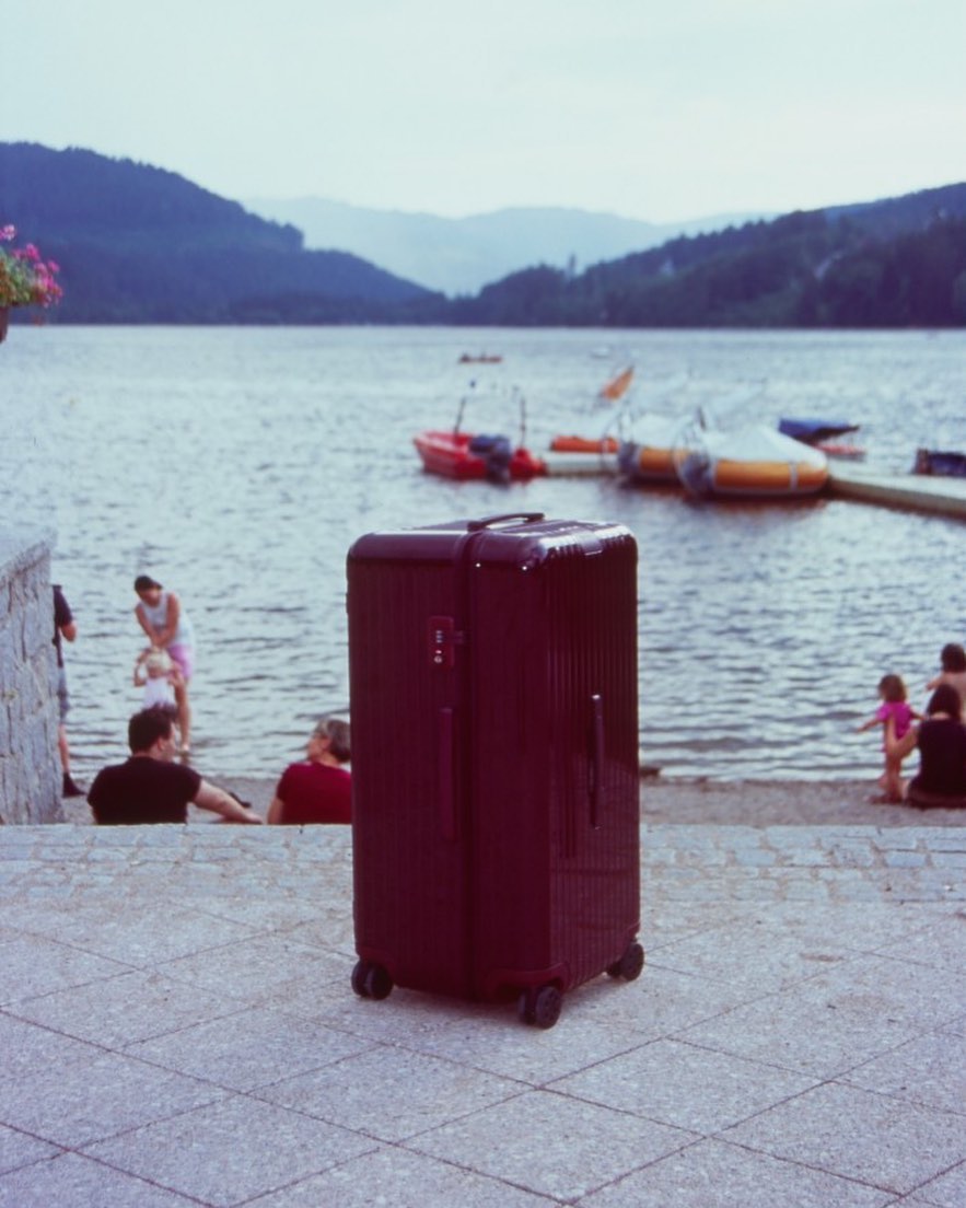 Maroon suitcase on gray cobblestone steps near a lake with distant mountains and boats, creating a travel or leisure scene.