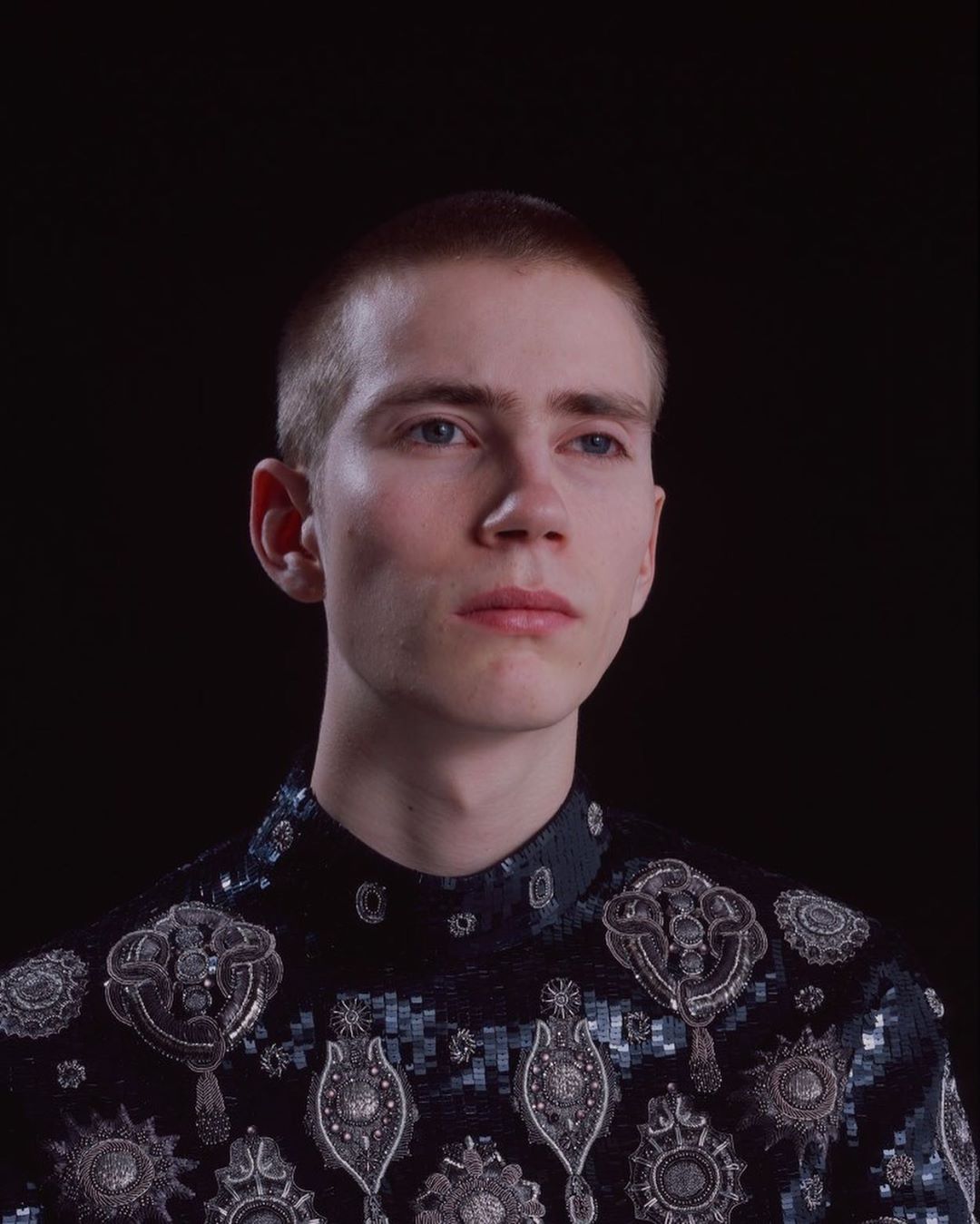 Close-up of a young adult male with a fair complexion wearing an ornate dark shirt against a stark black background in studio lighting.