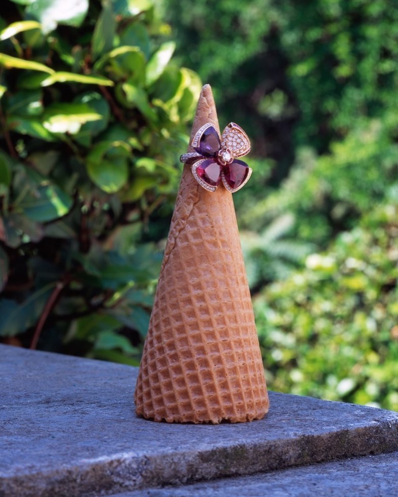 Flower-shaped ring with red and purple gems resting on top of a waffle ice cream cone, outdoors on a gray stone surface.