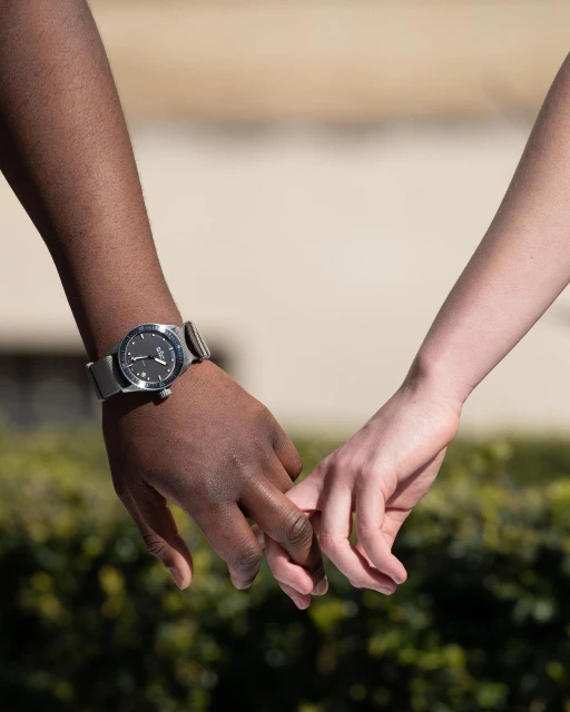 Interracial couple holds hands outdoors. Focus on hands and wrist watch. Close-up shot emphasizes connection and accessory detail.