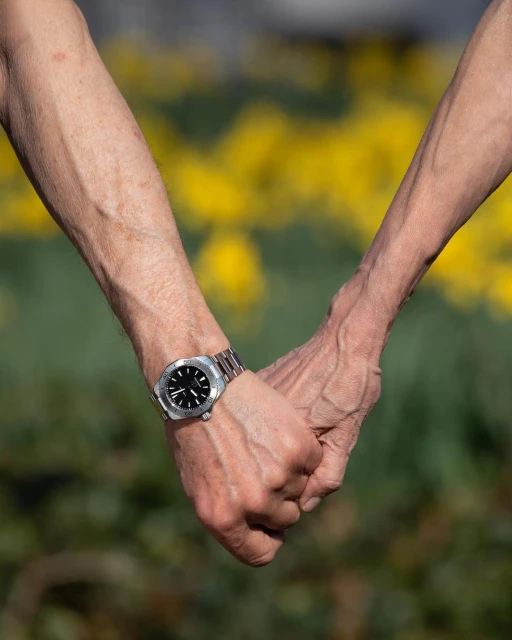 Two elderly hands clasped together wearing a wristwatch with blurred yellow flowers and green foliage in the background. 