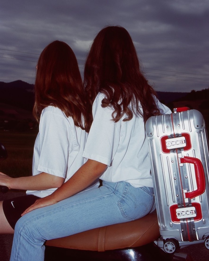 Two women with long brown hair, wearing casual clothes, sitting on a moped outdoors with a metal suitcase; mountains in the background.