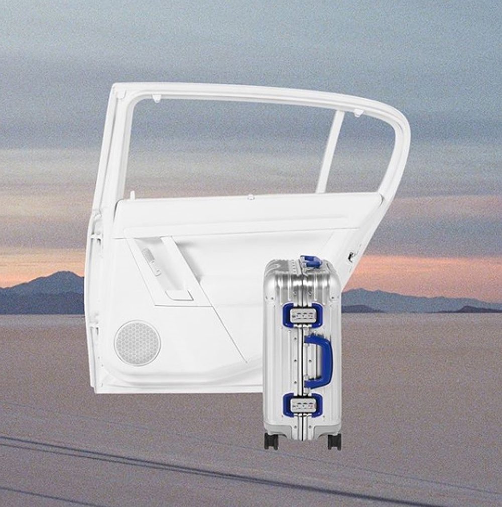 Silver rolling suitcase beside a white car door frame against a desert landscape with mountains and a colorful sky at dusk.