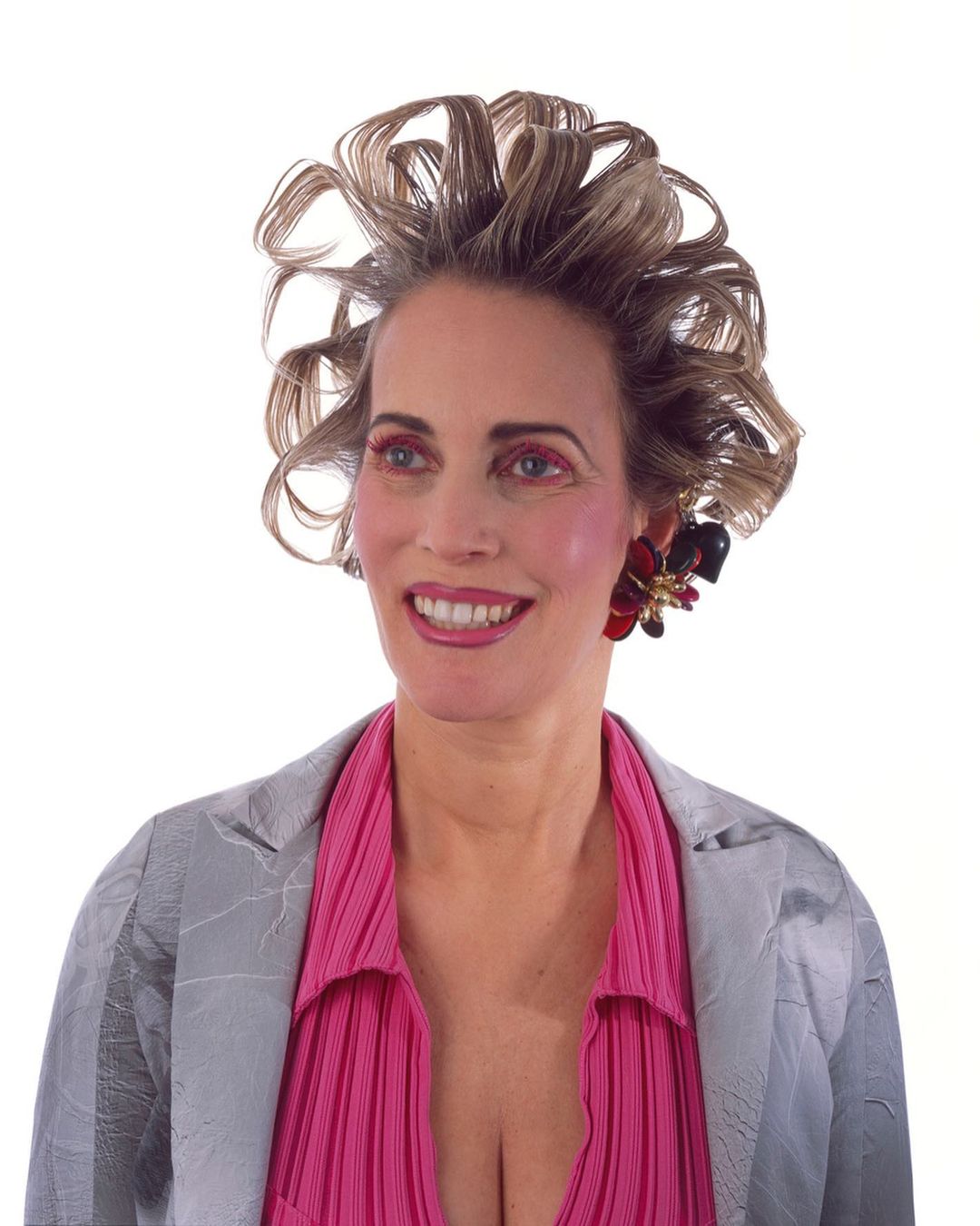 Smiling woman in a silver blazer and pink blouse against a white background with styled curly hair and floral earrings.