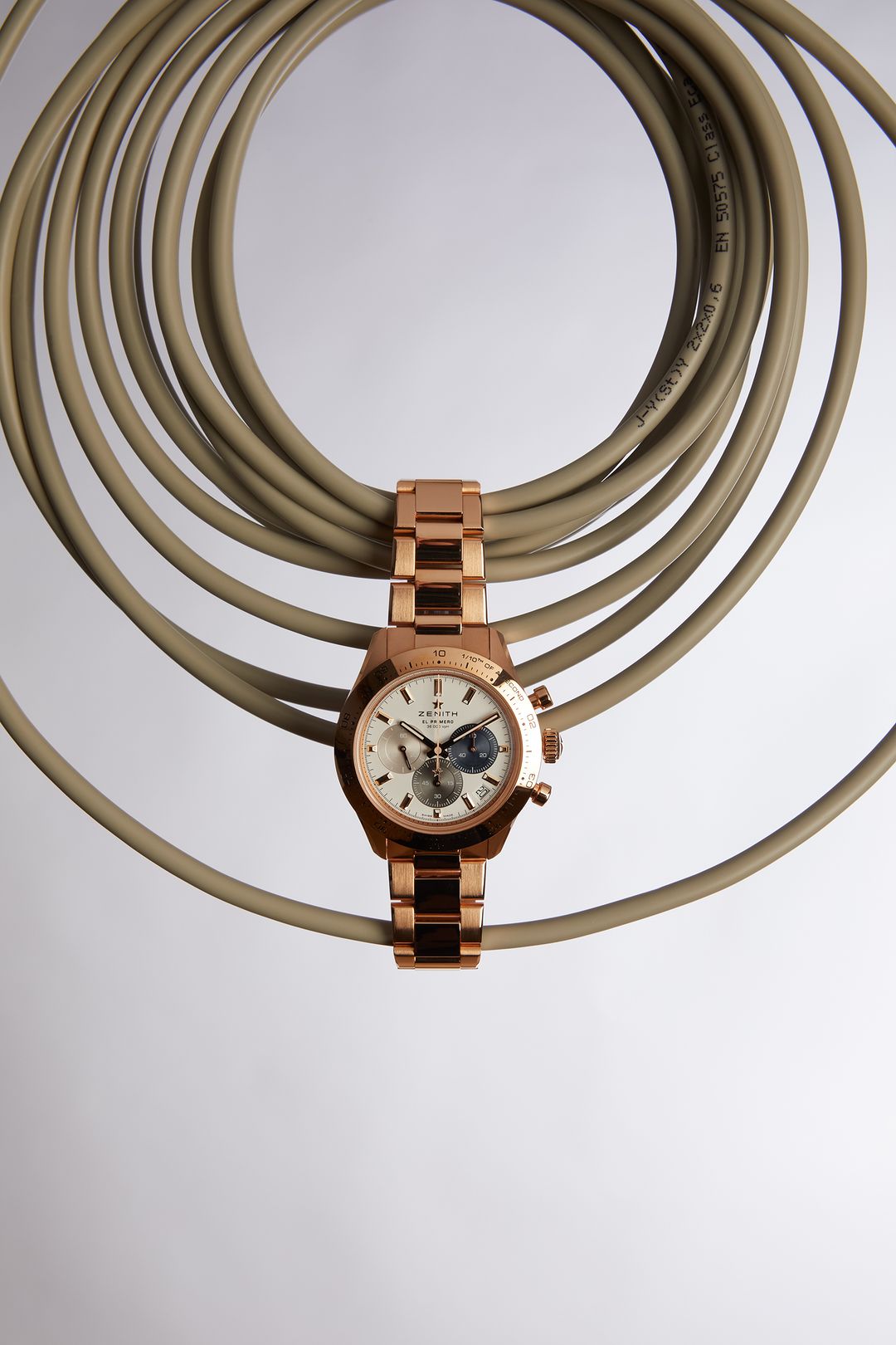 Rose gold Zenith watch with an open dial laying across a looped beige cable against a gradient white to grey background.