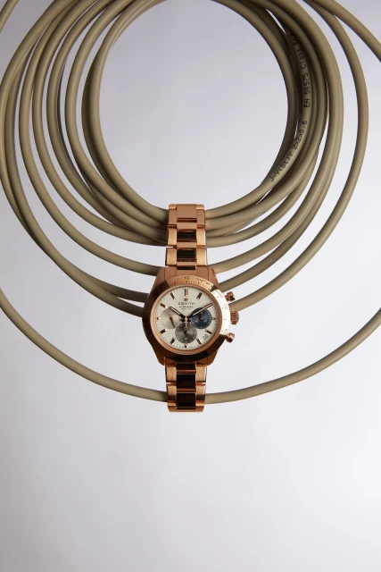 Rose gold Zenith watch with an open dial laying across a looped beige cable against a gradient white to grey background.