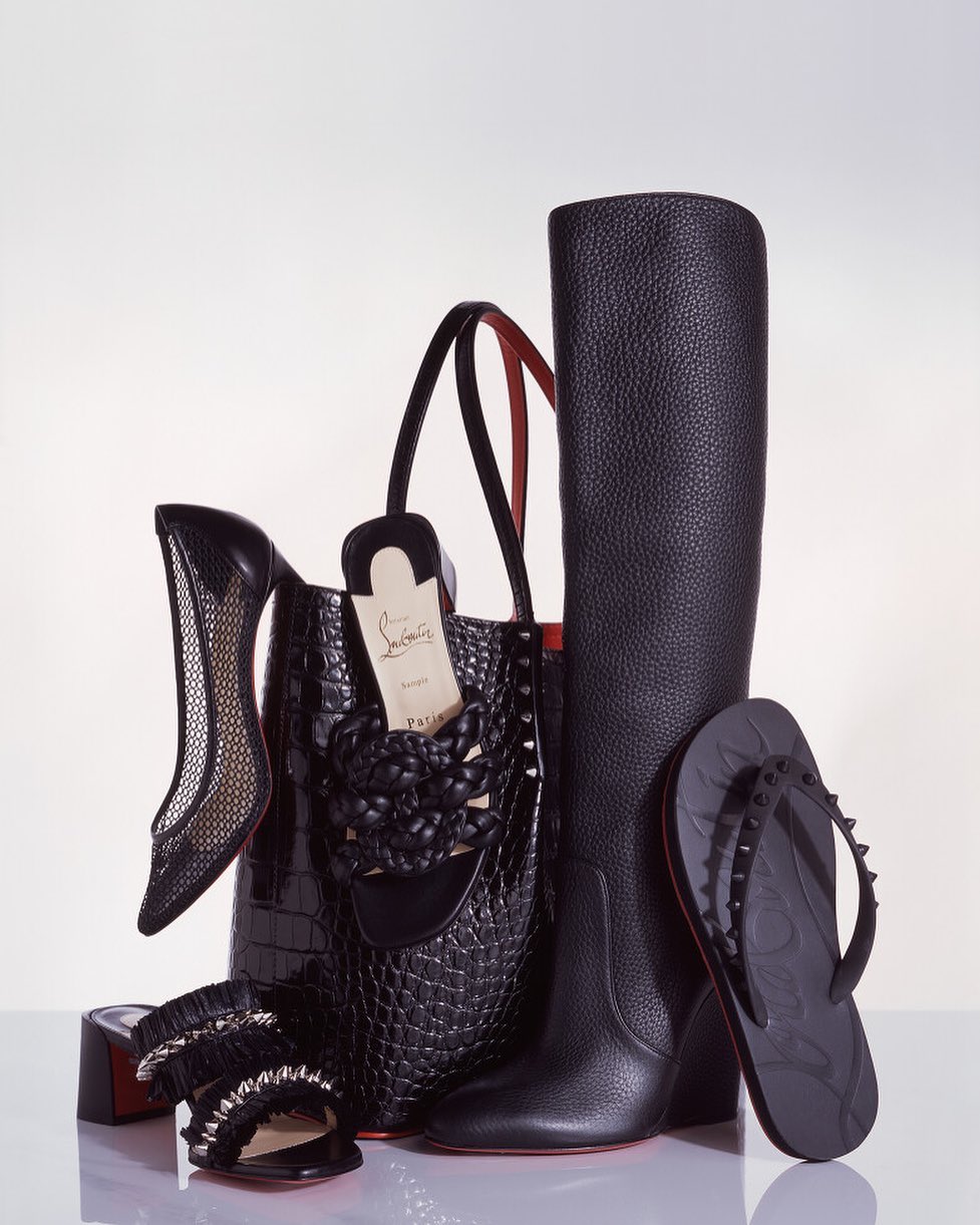 Arrangement of black Christian Louboutin shoes, boots, handbag, and sandal on a white surface, showcasing luxury fashion accessories.