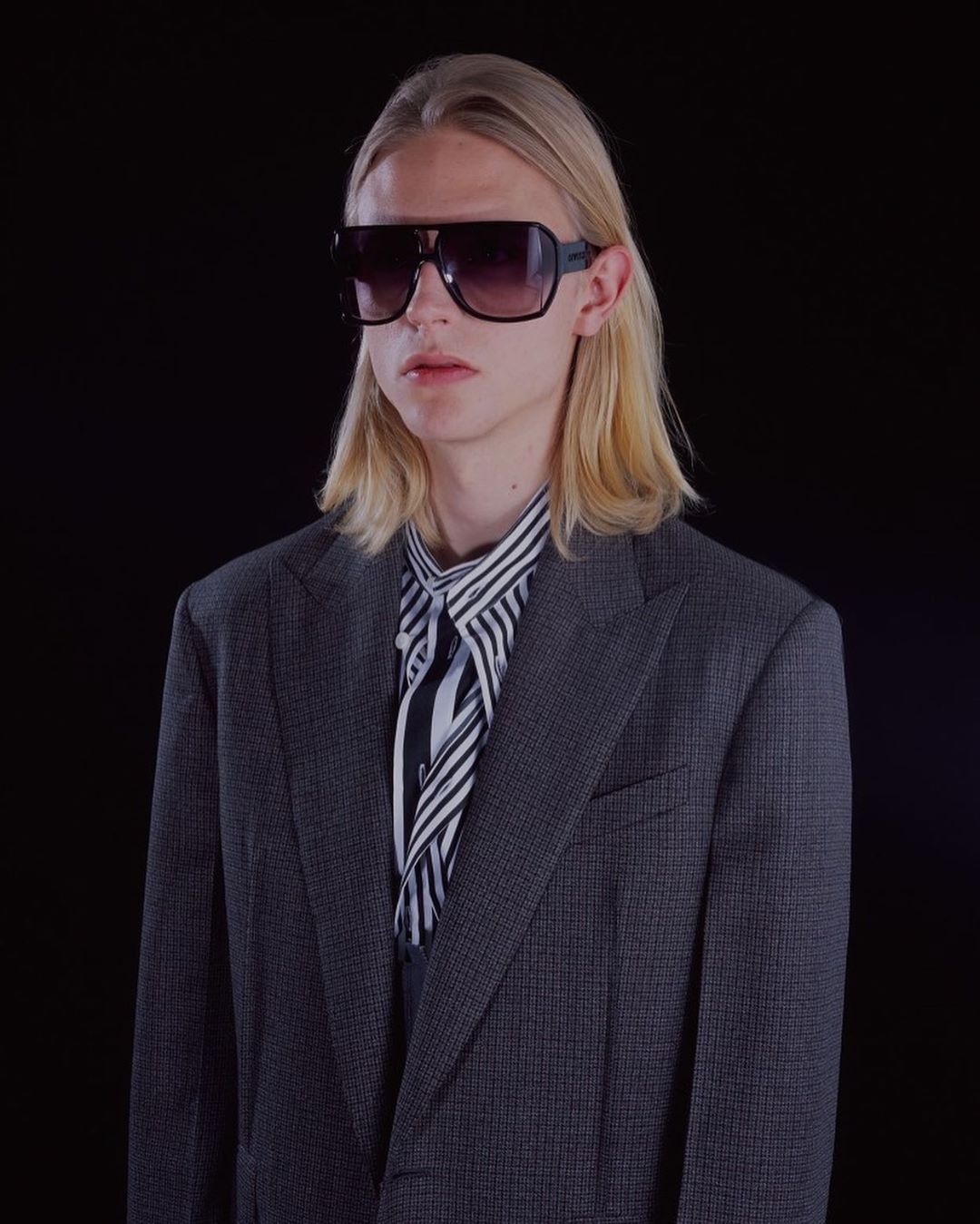 Adult man with blond hair wearing sunglasses, striped shirt, and grey blazer against a black backdrop in a studio shot.