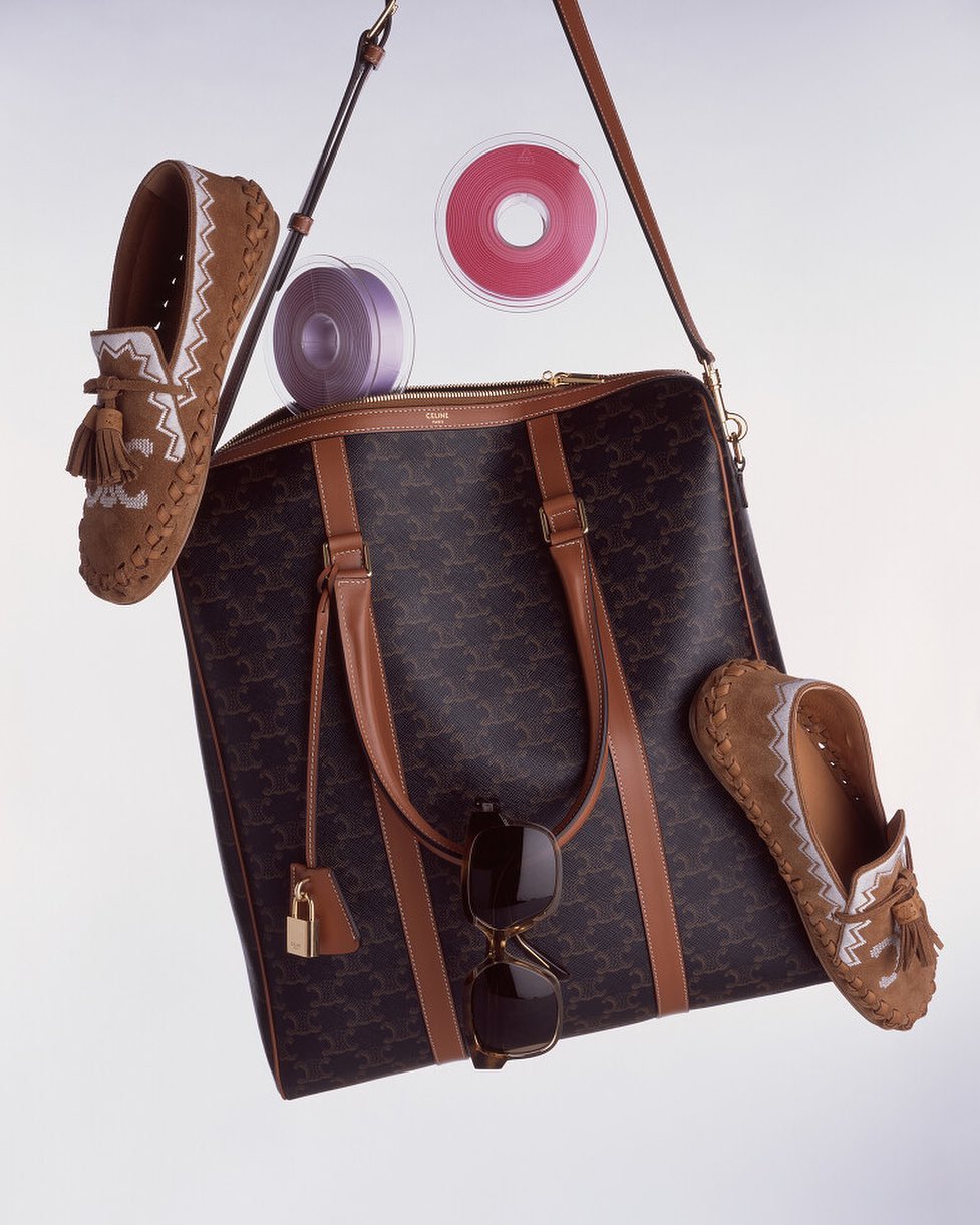 Luxury tan leather duffle bag with monogram, tassel loafers, ribbons, and sunglasses in a studio product shot for fashion editorial.