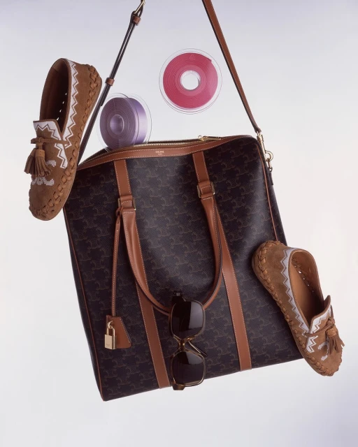 Luxury tan leather duffle bag with monogram, tassel loafers, ribbons, and sunglasses in a studio product shot for fashion editorial.