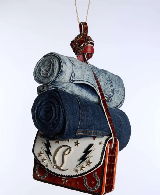 Stacked denim jeans and handbag with western-inspired design, suspended from a brown leather strap on a plain white backdrop.