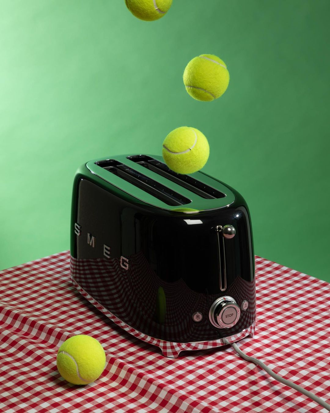 A black toaster sits on a red-and-white checkered tablecloth with tennis balls falling above it on a solid green background.