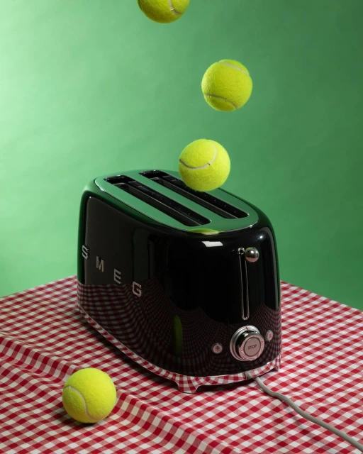 A black toaster sits on a red-and-white checkered tablecloth with tennis balls falling above it on a solid green background.