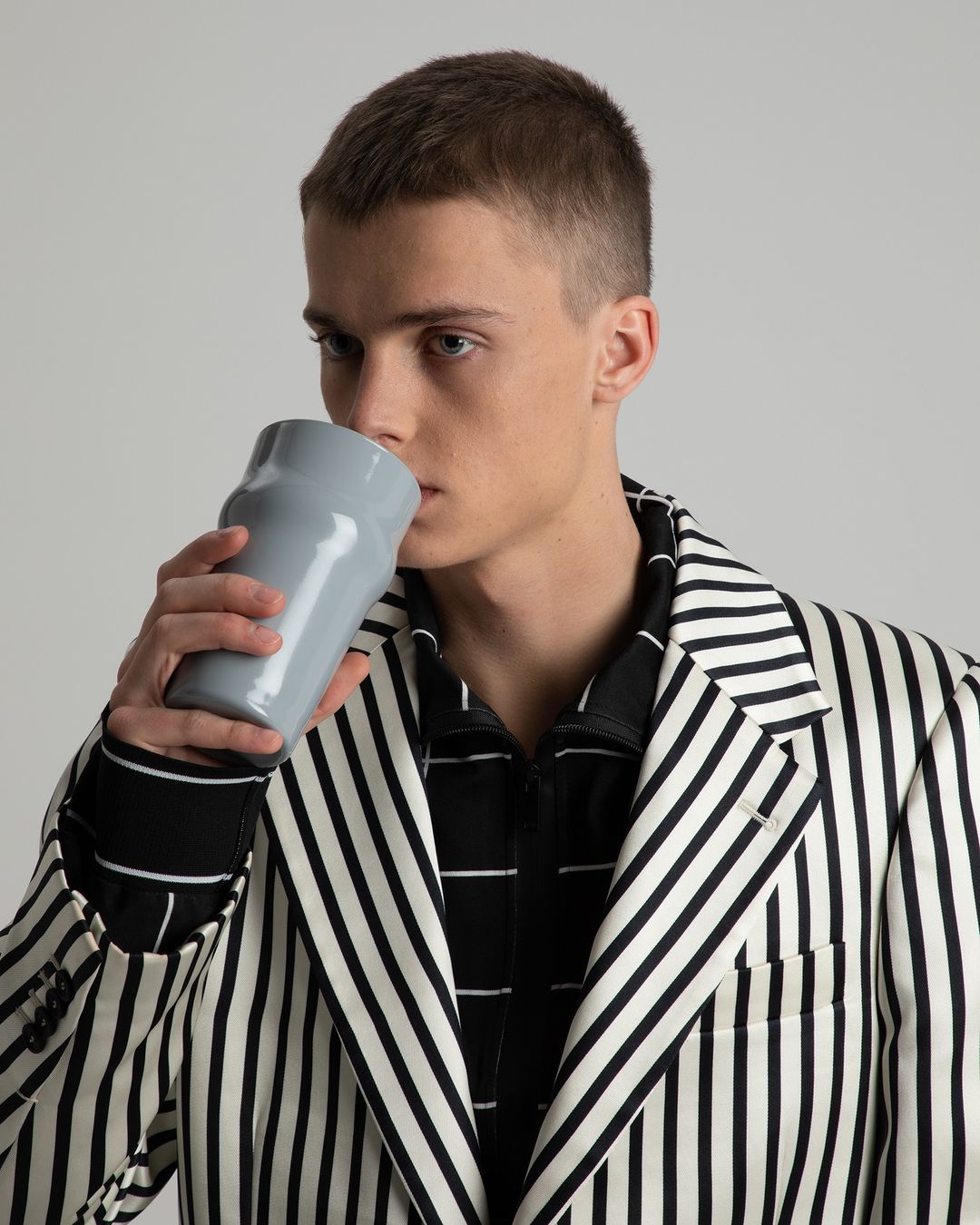 Young man in striped blazer drinks from grey ceramic mug, indoor studio lighting, neutral backdrop, wearing striped shirt.