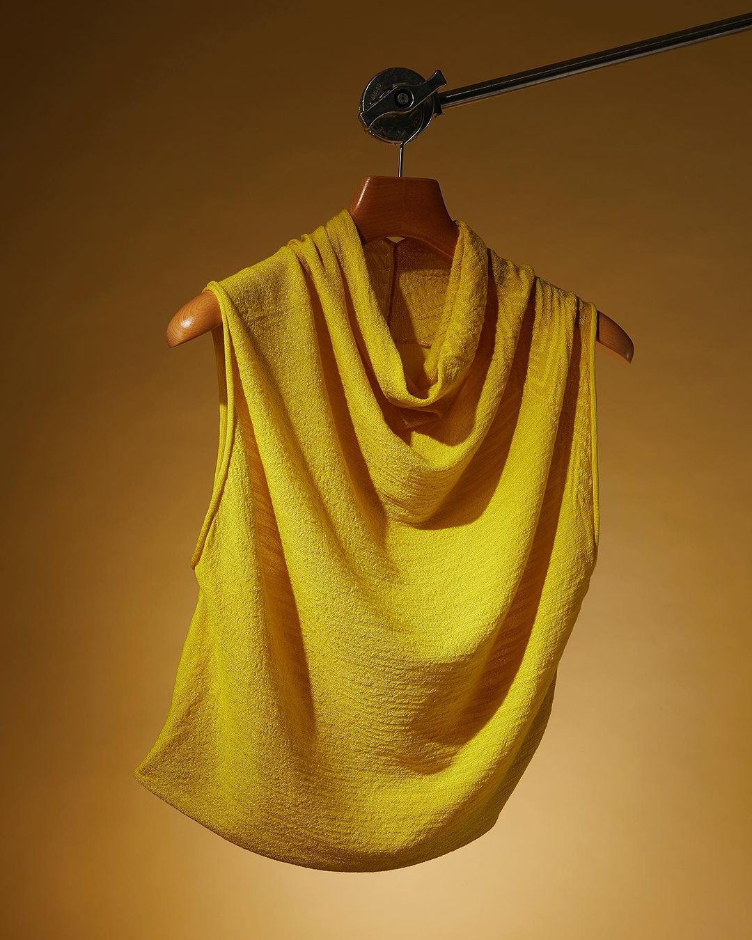 Yellow cowl neck top on wooden hanger. Sleeveless knit top with a draped neckline and textured fabric hanging against a tan background.