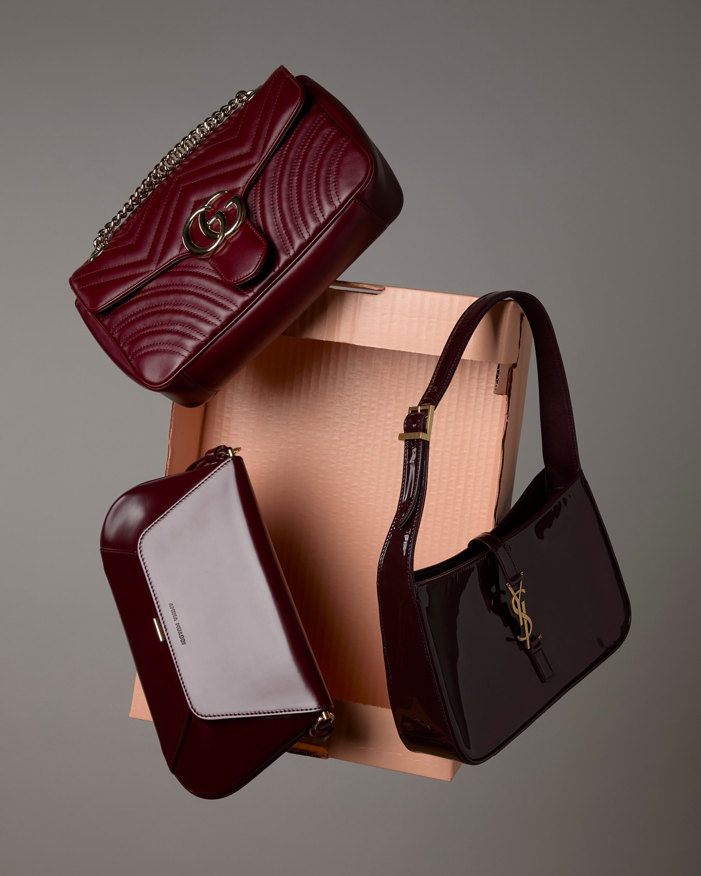 Three burgundy purses, including Gucci and Saint Laurent, are suspended in front of a light brown box and a gray background.
