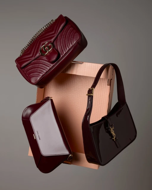 Three burgundy purses, including Gucci and Saint Laurent, are suspended in front of a light brown box and a gray background.