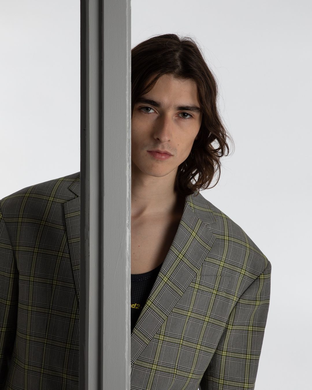A young adult male model with dark hair, wearing a grey and yellow plaid blazer, poses in front of a neutral white background.