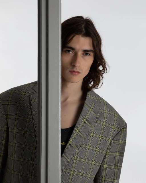 A young adult male model with dark hair, wearing a grey and yellow plaid blazer, poses in front of a neutral white background.