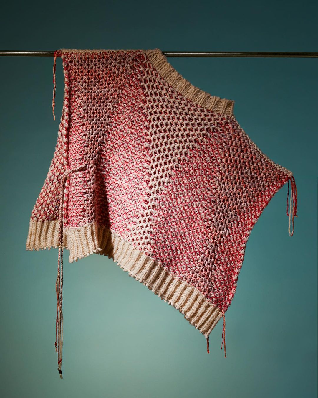 A knitted poncho in shades of red and beige is suspended from a metal bar against a teal backdrop, showcasing its open-weave design.