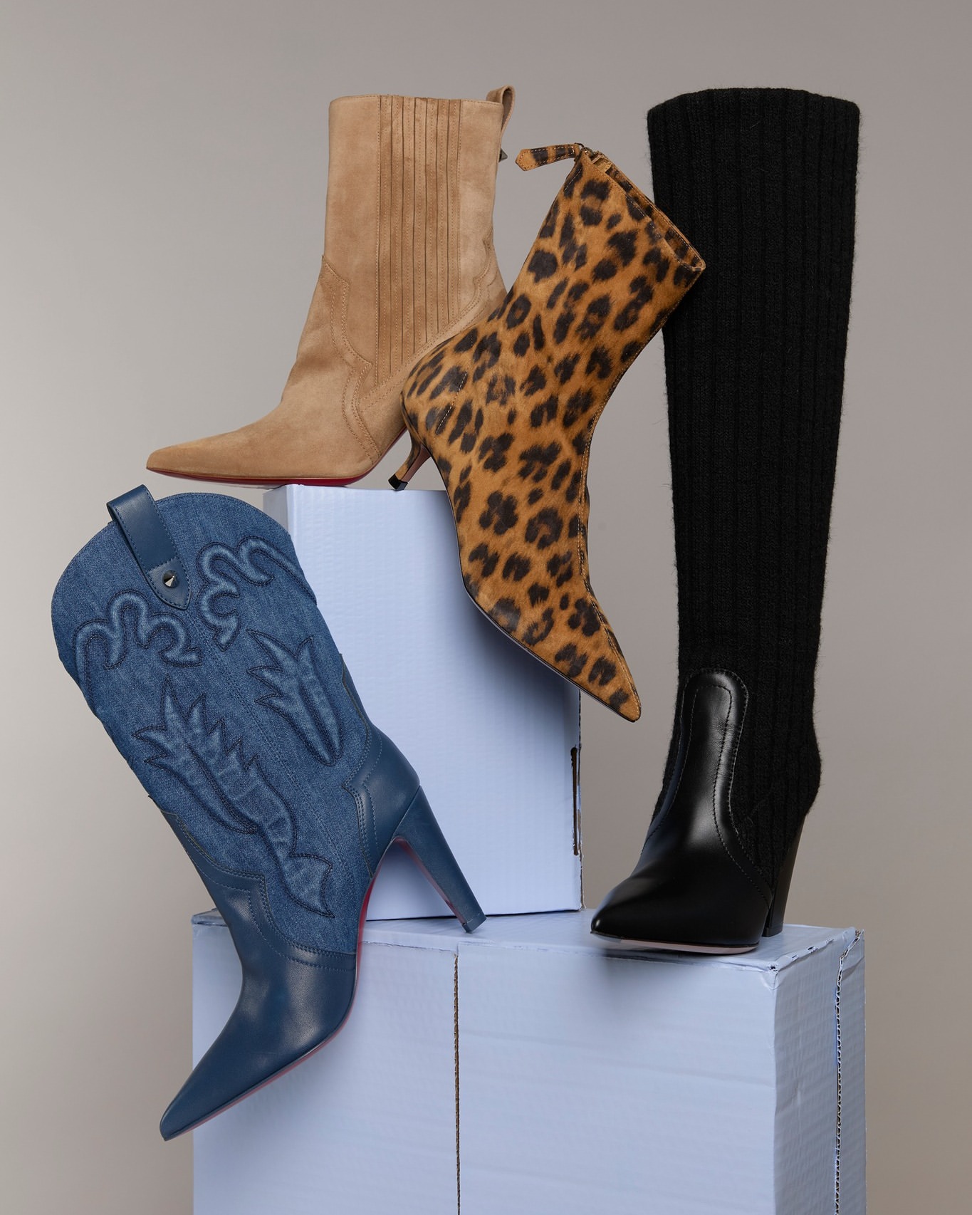 Four stylish women's heeled boots in different designs and materials including suede, leather, and animal print on stacked boxes.