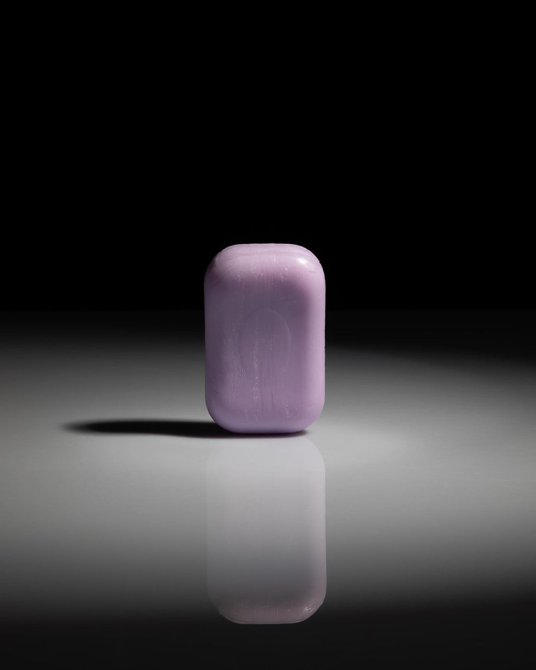 Studio shot of a pale purple rounded rectangular object on a gray surface with a dark shadow against a dark background.