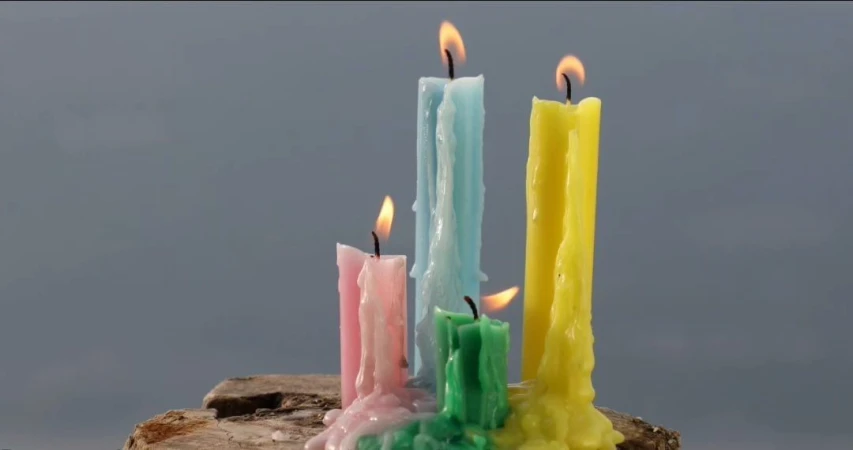 Group of lit candles in assorted colors on a wood surface against a plain background. Wax drips down the sides of each candle.