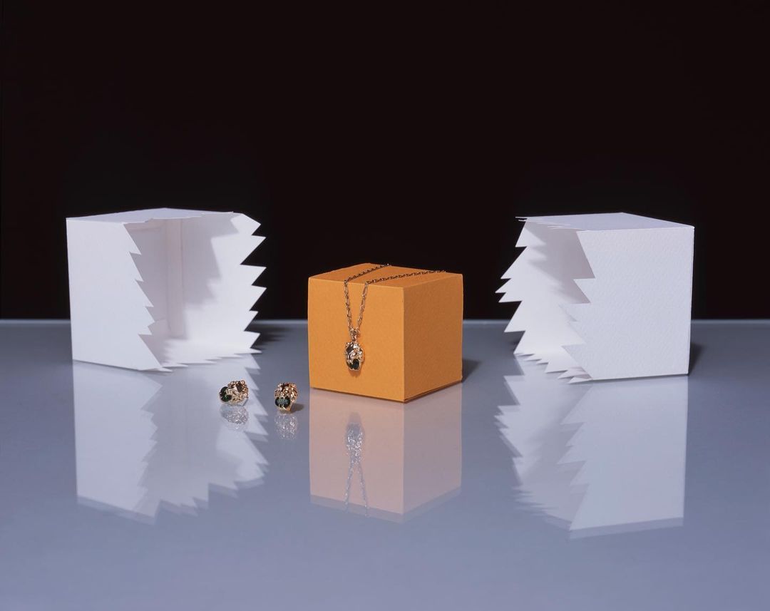 Jewelry still life: gold necklace on orange cube, with earrings and white geometric shapes against a dark backdrop, showcasing luxury.