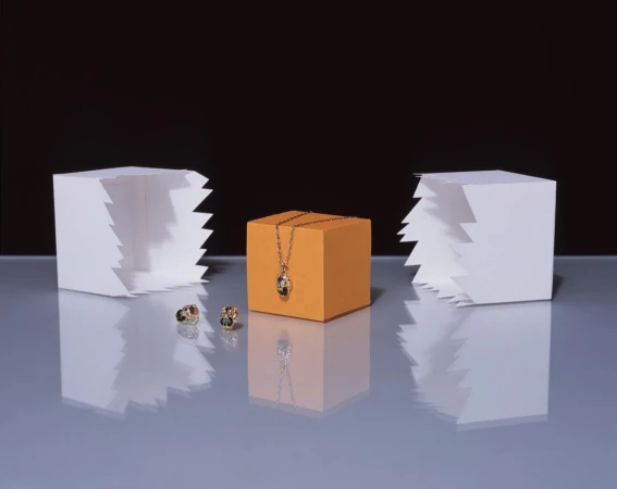 Jewelry still life: gold necklace on orange cube, with earrings and white geometric shapes against a dark backdrop, showcasing luxury.
