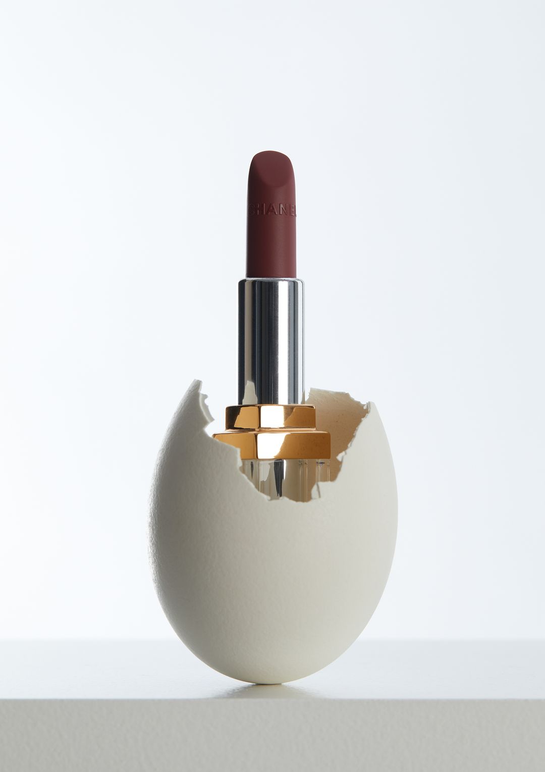 A lipstick standing in a cracked eggshell against a gradient white backdrop in a beauty advertisement concept.