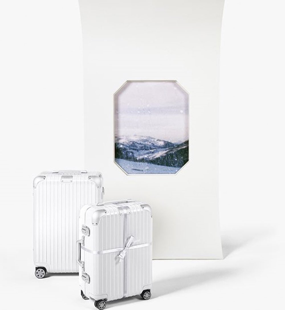 Two white ribbed suitcases sit next to an arched doorway with a snowy mountain landscape view in a minimalist, bright studio setting.