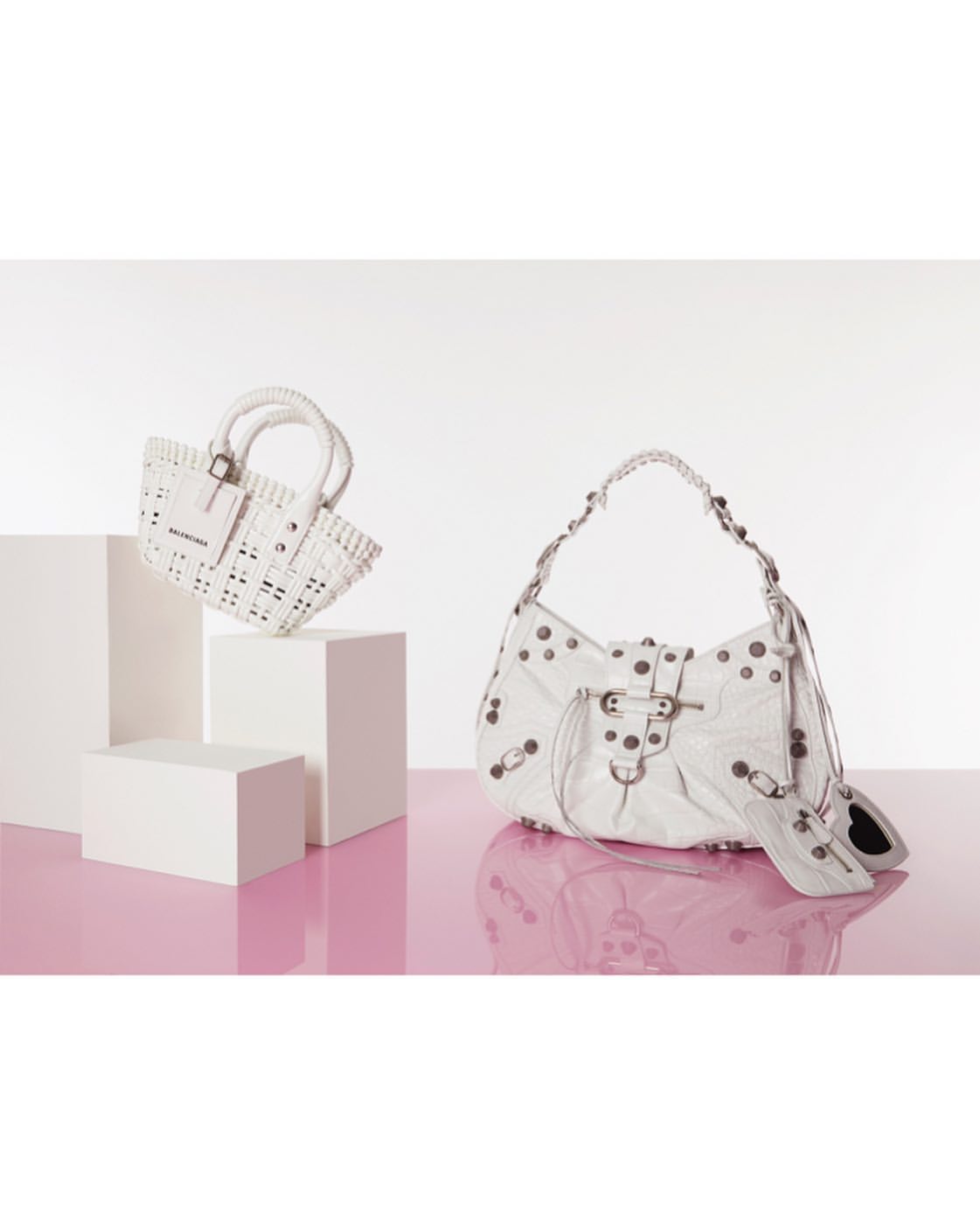 Two white Balenciaga handbags on white pedestals against a white and pink background, showcasing fashion and design elements.