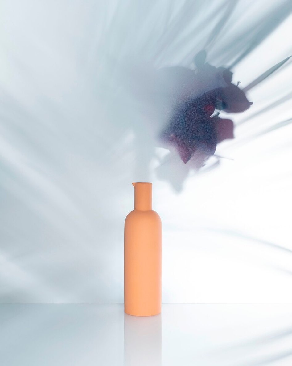 Minimalist product shot: a peach-colored bottle against a white background with floral shadows creating a soft, abstract aesthetic.