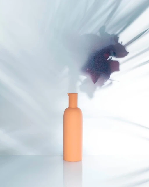 Minimalist product shot: a peach-colored bottle against a white background with floral shadows creating a soft, abstract aesthetic.