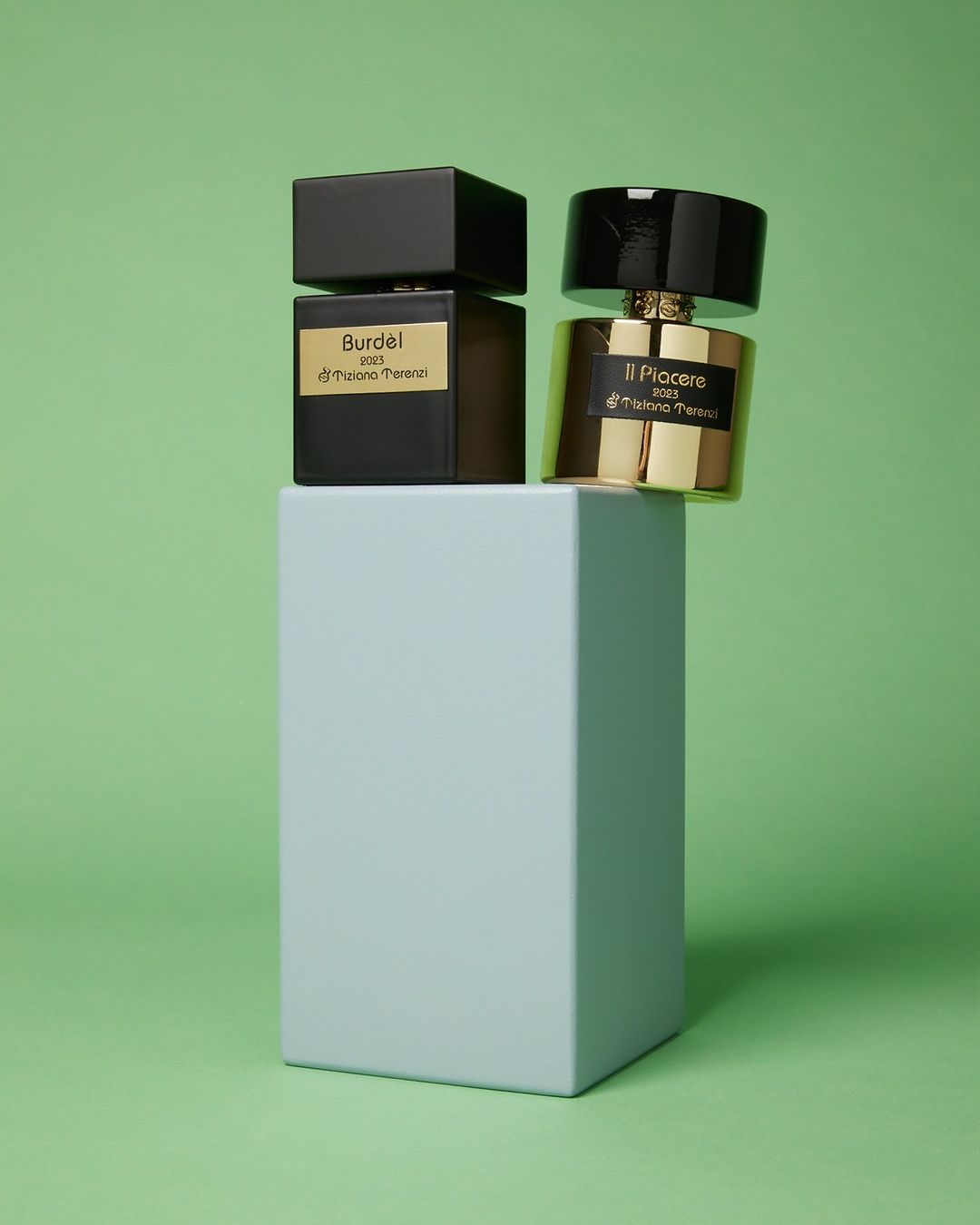 Two perfume bottles, one black and one gold, are displayed on a light blue pedestal against a light green backdrop in a product shot.