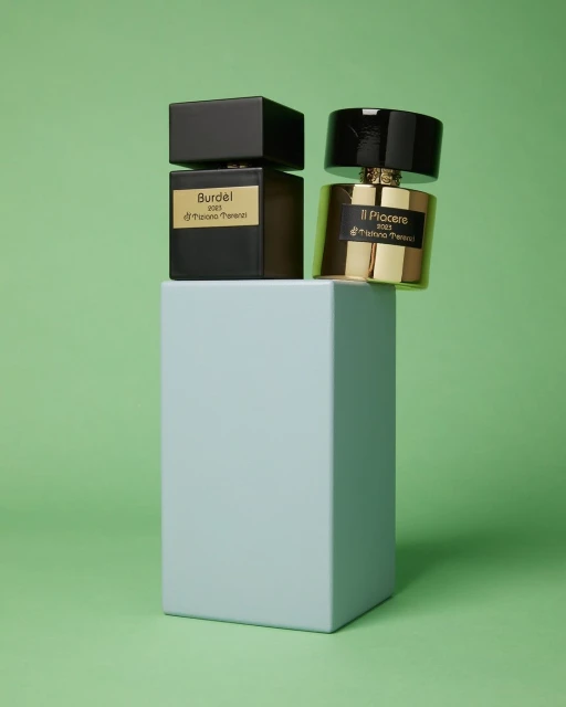 Two perfume bottles, one black and one gold, are displayed on a light blue pedestal against a light green backdrop in a product shot.