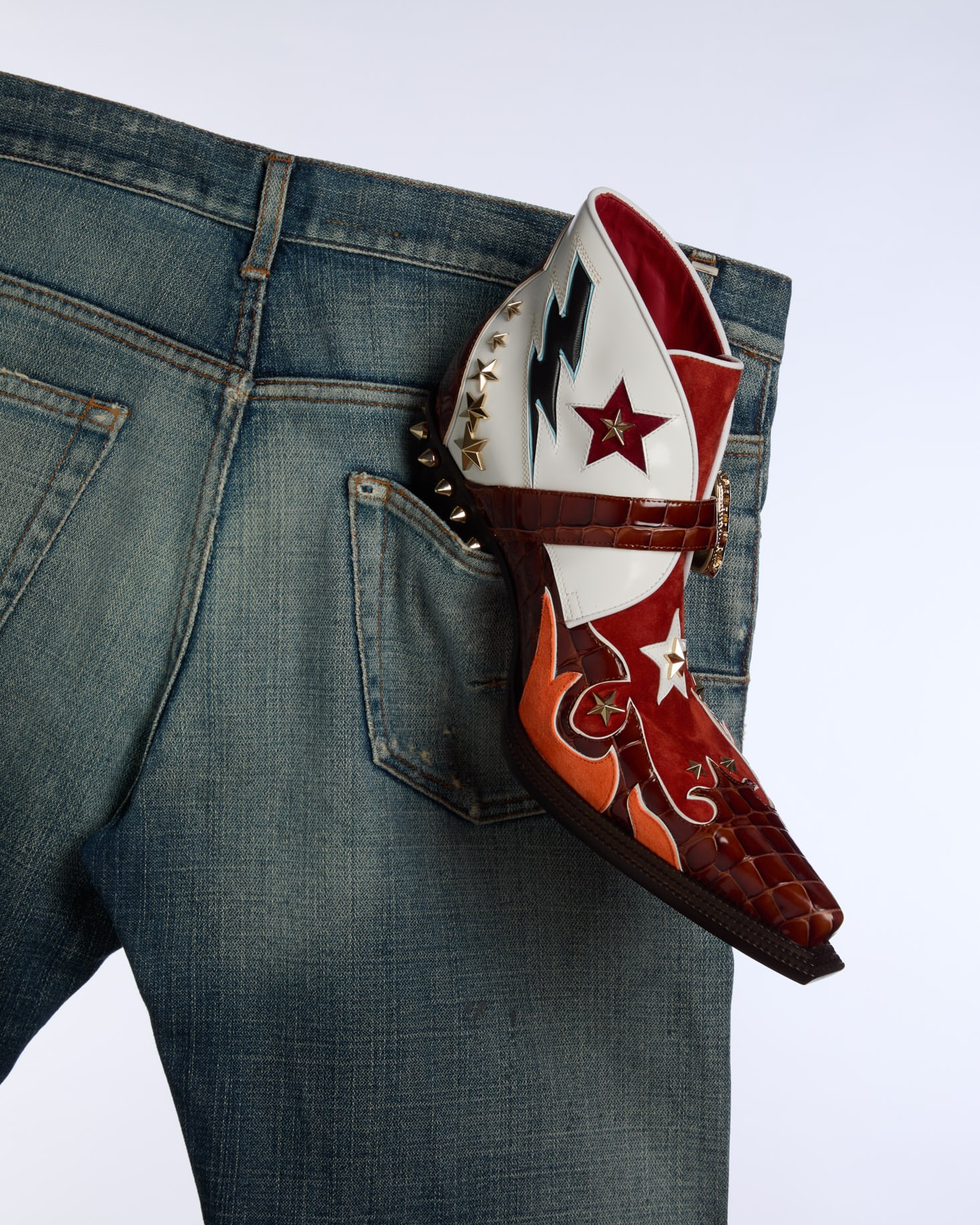 Denim jean with a colorful, patterned heeled boot sticking out of the back pocket on a white background. Fashion product shot.