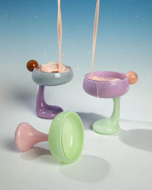 Three decorative pastel-colored glass stemware with milk-like liquid being poured into two glasses against a graduated blue background.